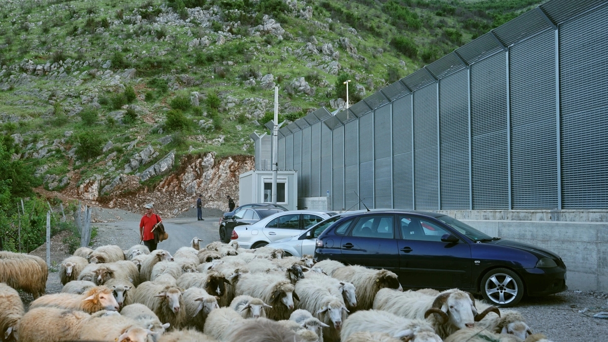 An Albanian shepherd drives his flock past the high wall of an EU migrant camp